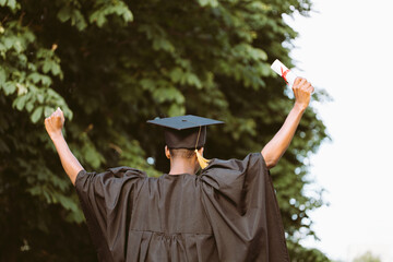 Afro american graduate from university in black mantle and hat stand with back outdoors among trees...
