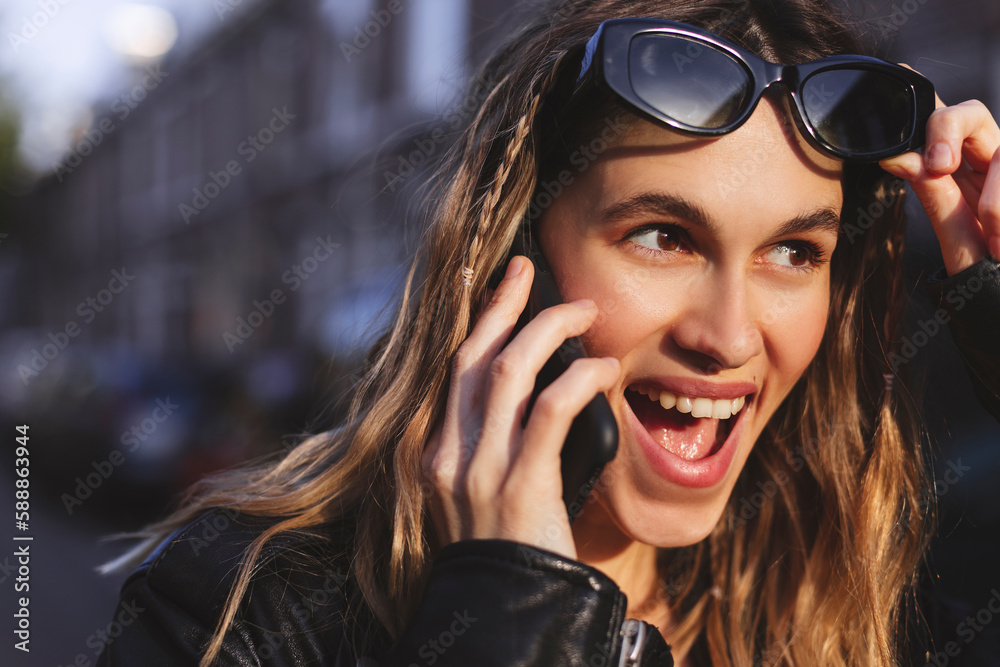 Wall mural Attractive happy woman talking on cell phone, raised her glasses in surprise, open mouth. Young cute tourist blonde woman with two thin braids outside, wearing black leather jacket and black dress.