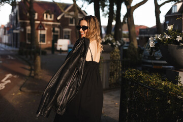 Pensive blonde woman wear black dress, glasses, leather jacket and touch it, posing. Outdoor shot of happy hippie lady with two thin braids and wave hair turn around. Girl hold jacket freedom style.