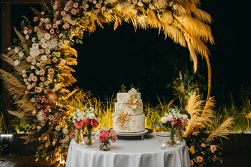 Three layered wedding cake at night, decorated with flowers made of frosting, on a round arch...