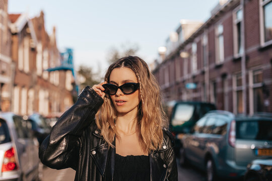 Pensive blonde woman in black leather jacket touch black glasses and posing on street background. Outdoor shot of happy hippie hipster lady with two thin braids and wave hair. Boho freedom style.