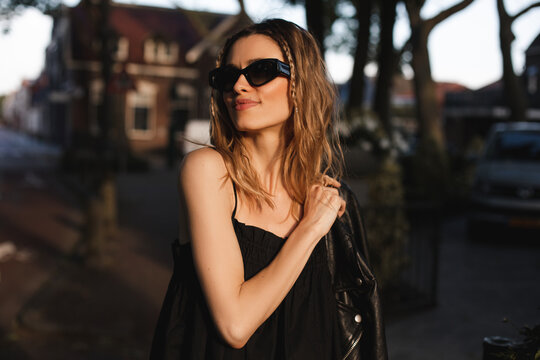 Pensive blonde woman wear black dress, glasses, leather jacket and touch it, posing, look side. Outdoor shot of happy hippie lady with two thin braids and wave hair. Coachella or boho freedom style.