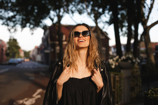 Pensive blonde woman wear black dress, glasses, leather jacket and touch it, posing. Outdoor shot of happy hippie lady with two thin braids and wave hair. Coachella or boho freedom style.