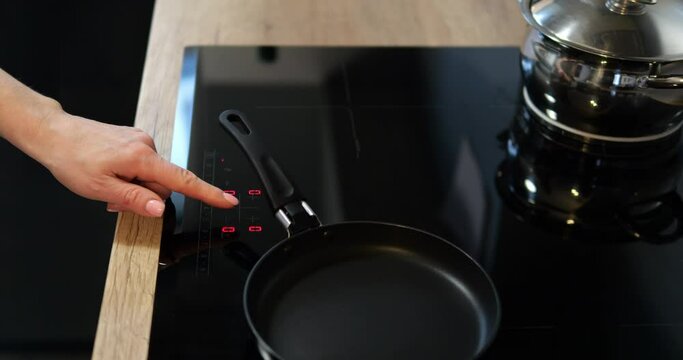 Female hand turn on black induction or electrical cooktop with pans. Comfortable kitchen equipped with modern convenient cooking device.