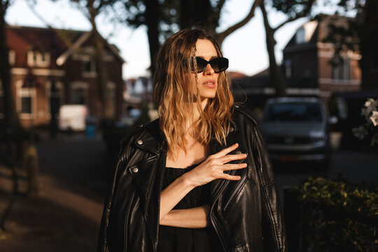 Pensive blonde woman wear black dress, glasses, leather jacket and touch it, posing. Outdoor shot of calm sensual hippie lady with two thin braids and wave hair. Coachella or boho freedom style.