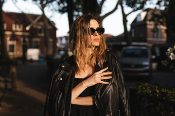 Obraz premium Pensive blonde woman wear black dress, glasses, leather jacket and touch it, posing. Outdoor shot of calm sensual hippie lady with two thin braids and wave hair. Coachella or boho freedom style.