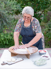 Front view of old female cook, chef wearing apron looking dowm , touching dough, rolling, making pita. Concept of fast food.