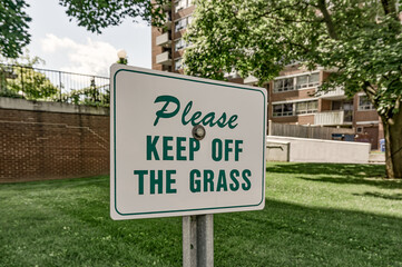 Fototapeta premium Please keep off the grass metal sign ,white background, green lettering, is on a metal pole outside a residential apartment building on a sunny summer day with green grass
