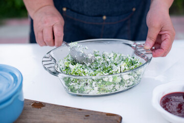 Close up of old female cook working outdoor in summer, holding spoon, mixing salad with cheese. Concept of cooking.