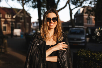 Pensive blonde woman wear black dress, glasses, leather jacket and touch it, posing. Outdoor shot of happy hippie lady with two thin braids and wave hair. Coachella or boho freedom style.