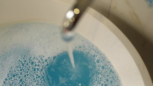Closeup top view of water running in bathtub with foam and colorful water after dissolve bath bomb. Preparation of taking bath at home or in hotel indoors. Concept of spa, relaxation after hard day.