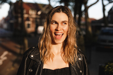Pensive blonde woman in black leather jacket and dress with open mouth posing on street. Outdoor shot of happy hippie lady with two thin braids and wave hair. Coachella or boho freedom style.