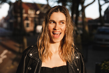 Fototapeta na wymiar Pensive blonde woman in black leather jacket and dress with open mouth posing on street. Outdoor shot of happy hippie lady with two thin braids and wave hair. Coachella or boho freedom style.