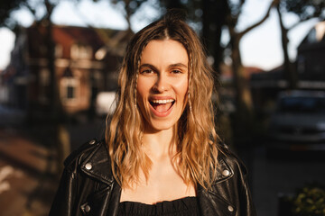 Pensive blonde woman in black leather jacket and dress with open mouth posing on street. Outdoor shot of happy hippie lady with two thin braids and wave hair. Coachella or boho freedom style.