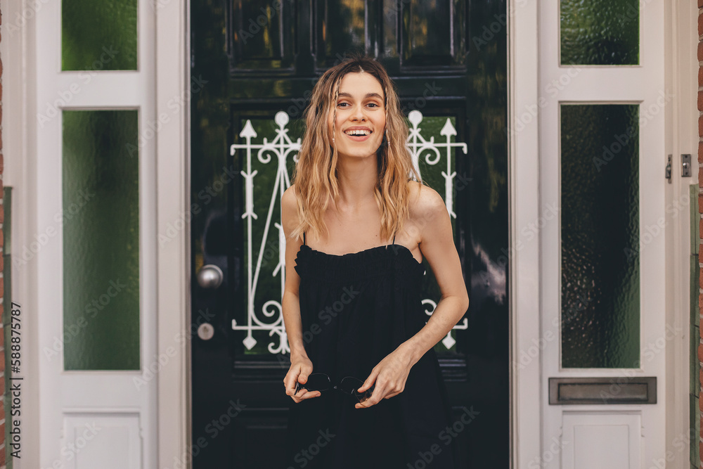 Wall mural Smiling young woman friendly cheerful smile to her new neighbours. Girl leaves the house closing the door and look happy, smiling. Girl wear black dress meeting friends. - Wall murals