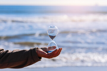 Time is running fast. Hourglass with blue sand inside in mature woman's hand symbolize the brevity of life. Background is sea with beautiful sunlights. Concept of the rapid passage of time. Copy space