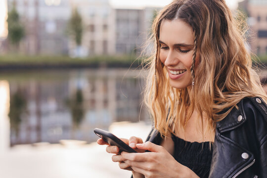 Happy blonde curly woman with two thin braids wear black leather jacket, dress hold her mobile phone and chatting with her boyfriend, girl smile when scrolling social media page or read good news
