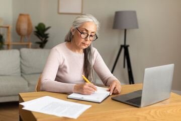 Concentrated serious busy pretty caucasian old lady in glasses makes notes at table with computer