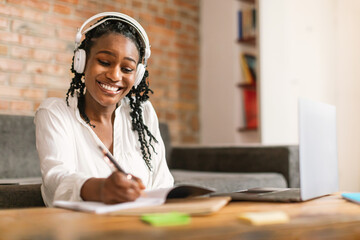 Happy black woman in headphones studing online with laptop from home, watching webinar and taking...