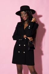 Fashionable young woman with black coat, hat and small bag. Stylish outfit on pink background.