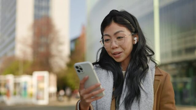 Attractive Asian Woman Standing Outside, Using Smartphone, Videocalling to Her Friends, Waving at the Camera Using Earphones. Lady Talking Online. People and Technology, Commucication Concept