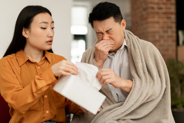 Asian middle aged man blowing nose in napkins, loving caring wife treating her ill husband, taking care