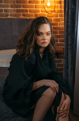 Portrait of a beautiful woman in a black bathrobe, who is sitting on the bed, looking at the camera