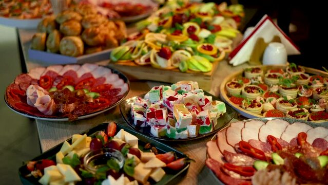fourchette table in a nightclub or restaurant with tasty delicious dishes,cheese asorti,fruits,spread with prosciutto,olive,jelly colorful cake.dark walls pour light condition 4k rotating spinning