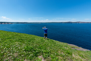 A senior Caucasian man standing wearing a blue shirt and green shorts looking out over the ocean on a sunny day. The calm ocean and clear sky are deep blue in color. The shoreline is covered in grass.