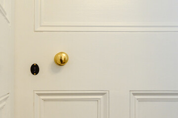 A vintage white color wooden interior panel house door with a small round gold doorknob and a brass keyhole. The door has boards, and molding and is heavily painted. 