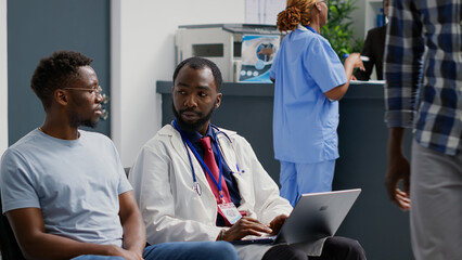 Obraz premium General practitioner using diagnosis and results on laptop to show medicare report to male patient. Doctor working with computer to explain disease treatment, consultation in hospital lobby.