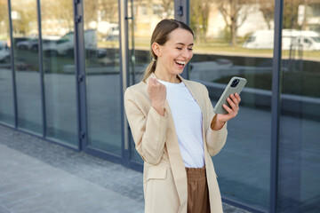 Fototapeta premium Excited businesswoman walking near office building and feeling surprised while looking on smartphone. Happy female worker making yes gesture after reading good news