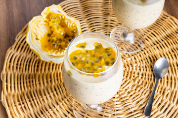 Passion fruit mousse in a glass bowl, with passion fruit in the background, on a straw disc and a wooden table. Detail silver spoon.