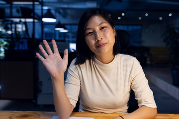 Happy asian businesswoman sitting and waving, making video call, working late at office