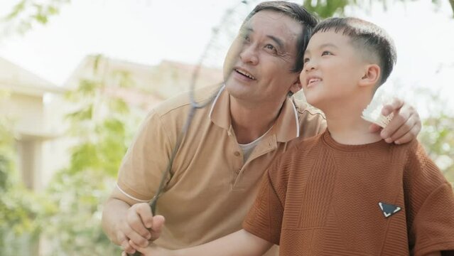 Tilt up shot of Asian father embracing son and showing him how to hold badminton racket outdoors in park on summer day