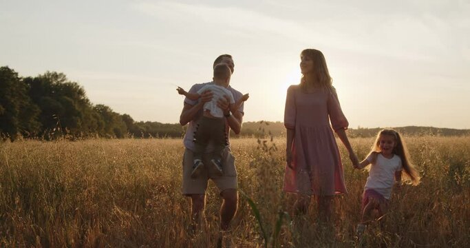 Happy young smiling family of four spend time in a golden wheat field at sunset. Mom, dad, son and daughter walk together during golden hour. Father plays with son throwing him up in the air.