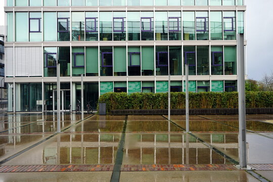 Heinz-Raspe-Platz mit gr&uuml;ner Hecke und moderner Architektur bei Regen an der Lindleystra&szlig;e und dem Main Riedgraben im Ostend von Frankfurt am Main in Hessen