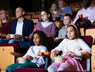 Portrait of tween girl with aframerican friend sitting in movie theater with popcorn and watching...