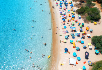 Aerial view of colorful umbrellas, sandy beach, swimming people in blue sea, trees at sunset in...