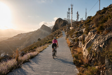 Fit female cyclist wearing cycling kit and helmet riding on the road on a gravel bike at sunset.Empty mountain road. Sports motivation image.Calpe town in Spain.