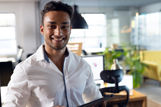 Portrait of happy biracial businessman smiling, holding tablet, working late at office