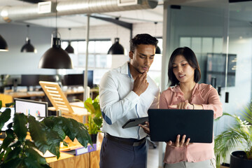 Diverse business people using tablet and laptop, discussing, working late at office