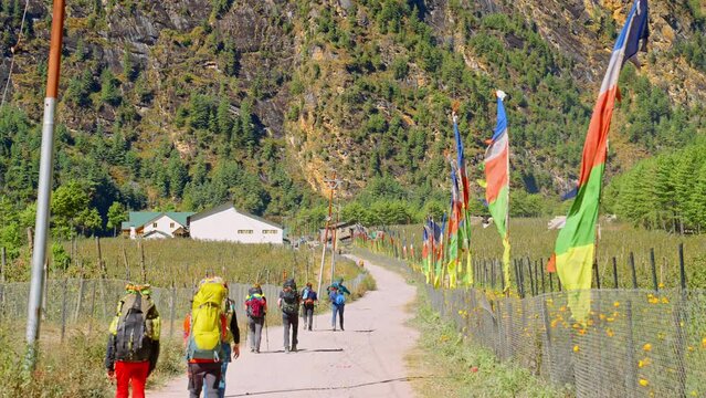 Following a group of backpacking hikers walk past rows of prayer flags towards steep Himalayan mountain cliff, Annapurna Circuit Trek, Nepal