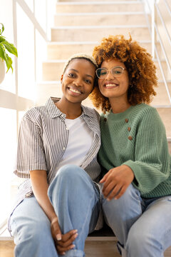 Portrait of multiracial young lesbian couple smiling and sitting on steps at home, copy space