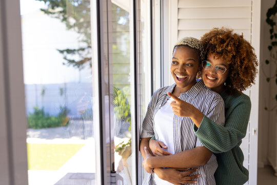 Cheerful multiracial young lesbian couple cuddling and looking through window while standing at home