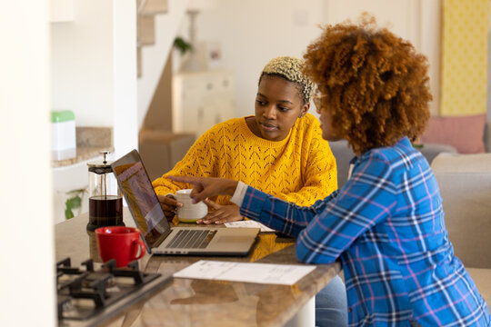 Diverse lesbian couple analyzing bills over laptop while having coffee on kitchen island