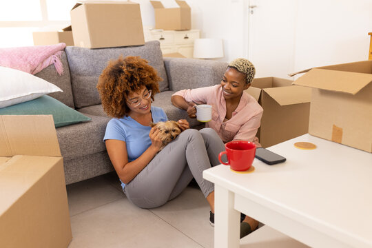 Multiracial lesbian couple with cairn terrier relaxing and having coffee around boxes in new home