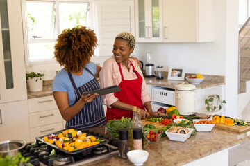 Happy diverse lesbian couple watching recipe over tablet and chopping vegetables on kitchen island