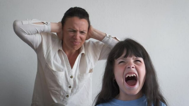 The child screams loudly against the background of the shocked mother. Portrait of an angry emotionally screaming little girl. Attention deficit hyperactivity disorder.