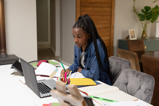 Happy african american teenager girl sitting by table, doing homework and using laptop - Powered by Adobe